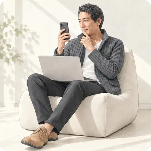 ソファでノートパソコンを膝に置き、スマートフォンの画面を興味深そうに見つめる男性。検索画面でAI Modeの答えに見入る様子をイメージ。