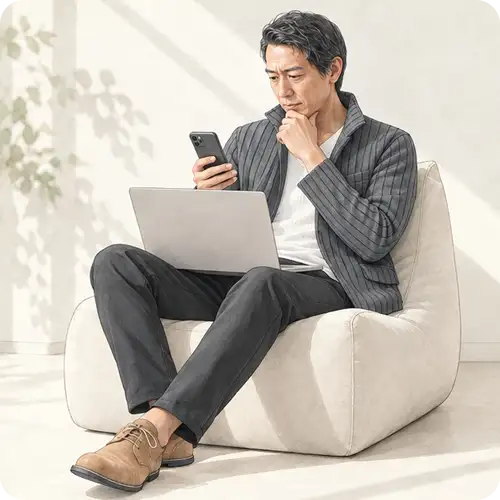 ソファでノートパソコンを膝に置き、スマートフォンを見ながら顎に手を当てて考え込む男性。Google広告のDSAからAI Maxへの移行を思案するイメージ。
